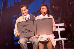 Two actors sitting on a stage read an oversized Romeo and Juliet book.